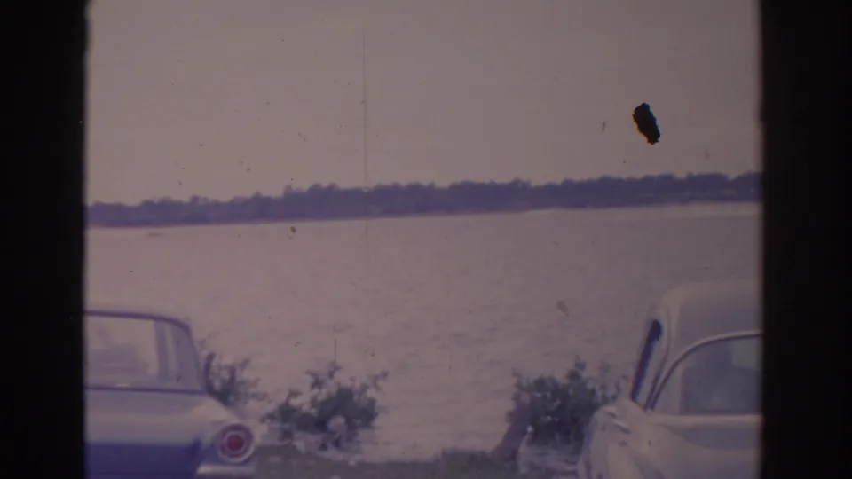 1962 Florida 8mm frame showing two cropped vintage cars near the lake, with a small distant boat and tree-lined horizon.