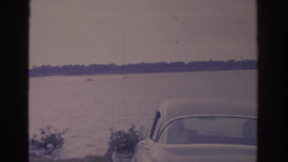 1962 Florida home-movie frame with a cropped vintage car in the foreground, lake water, distant boat, and low wooded shoreline.