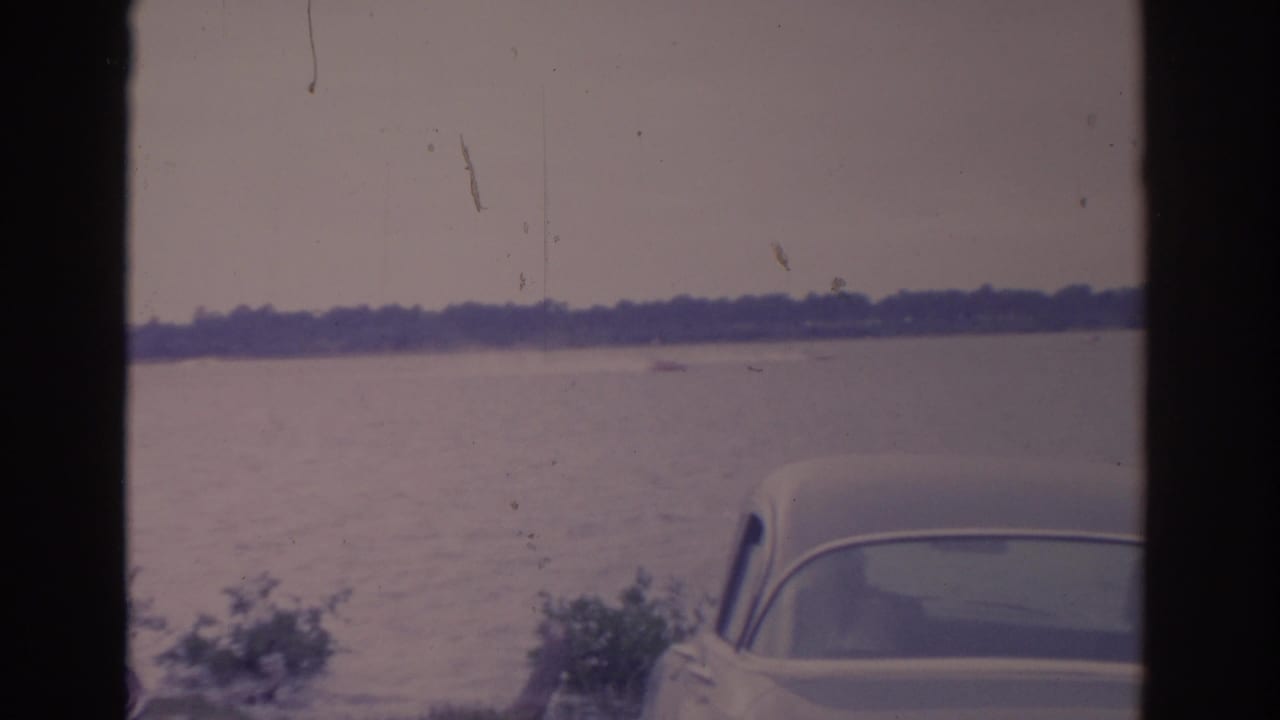White car parked near a calm lake with a distant shoreline and dark film gate edges.