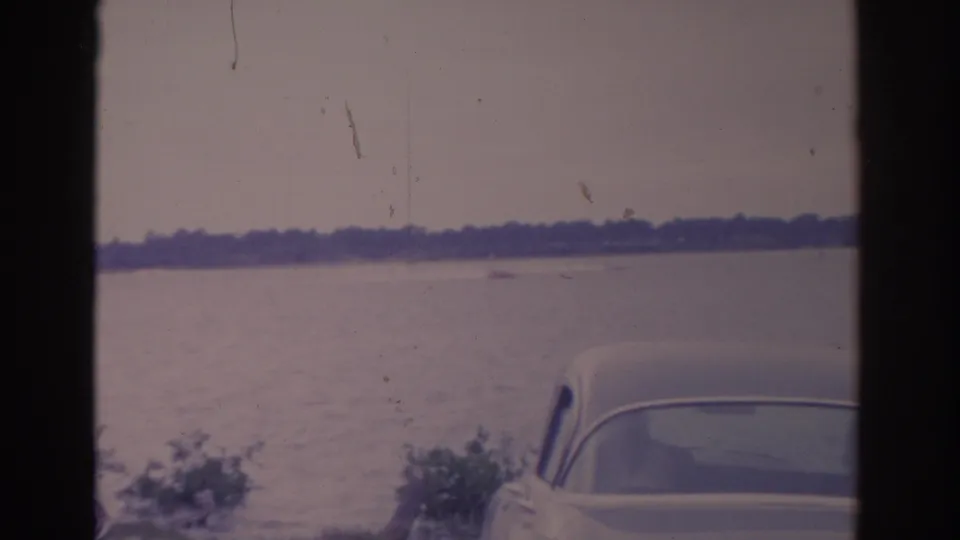 1962 Florida 8mm frame showing a white car by a broad body of water, with haze, distant trees, and visible film borders.