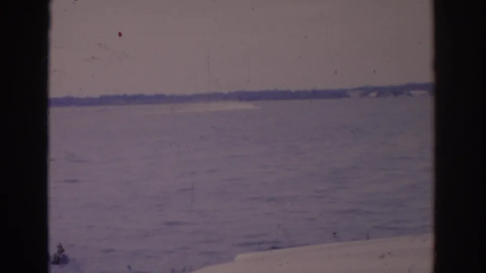 1962 Florida home-movie view across a broad lake, with a partial parked car in the foreground and a low distant shoreline.