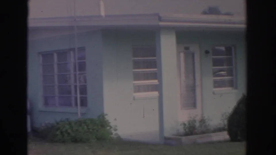 1962 Florida 8mm frame of a light green mid-century house exterior with jalousie-style windows, a screened door, shrubs, and lawn.