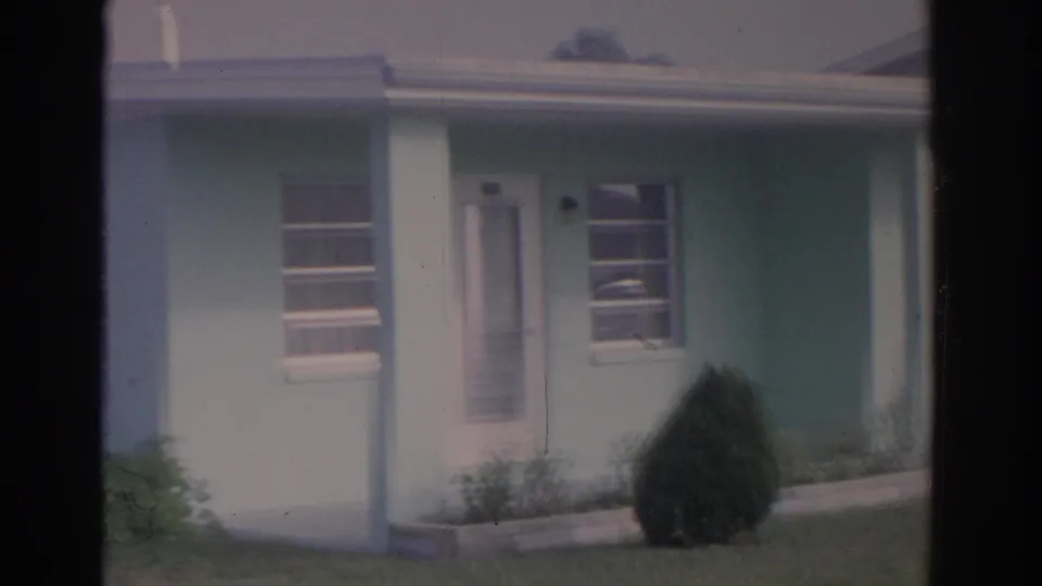 1962 Florida home-movie frame showing the front of a modest light green house with a screened door, windows, porch post, shrub, and lawn.