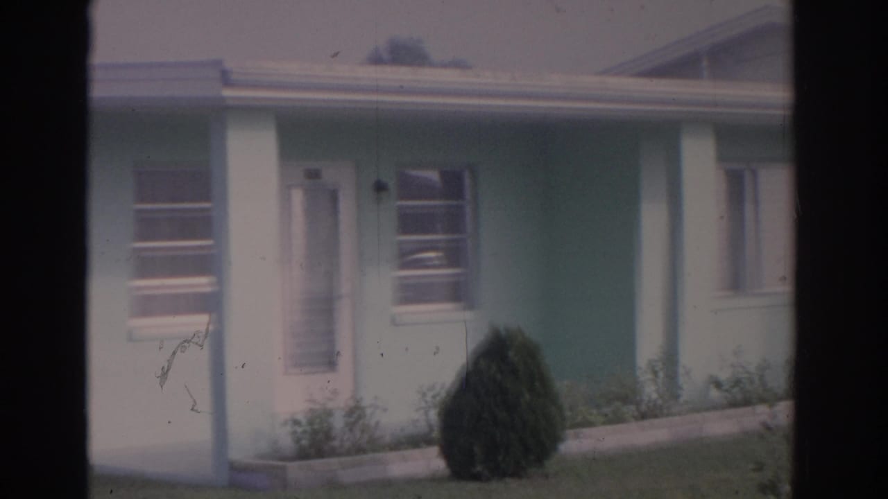 1962 Florida 8mm frame showing a light blue house with white trim, a front door, window, porch post, lawn, and conical shrub.