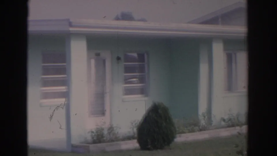 1962 Florida 8mm frame showing a light blue house with white trim, a front door, window, porch post, lawn, and conical shrub.