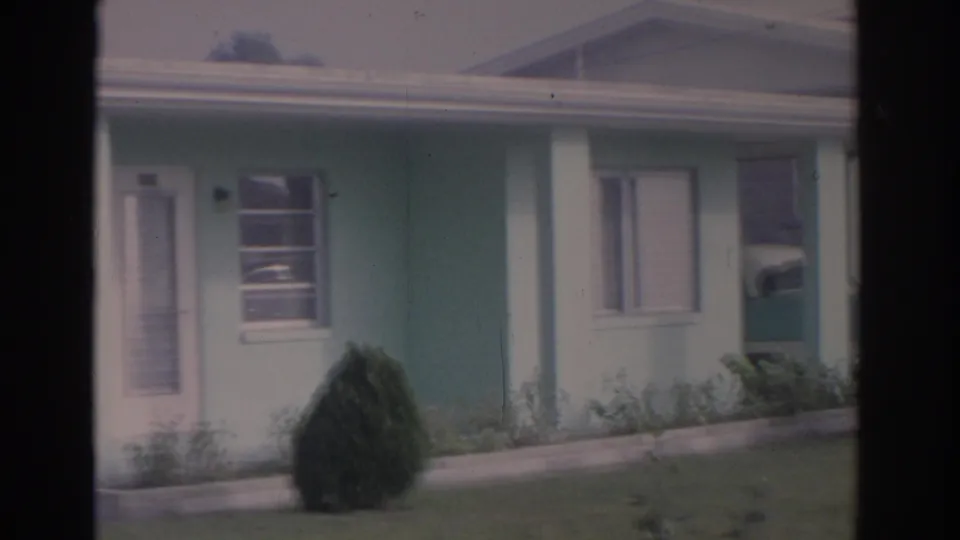 1962 Florida home-movie frame with the camera panning across the blue house facade, white porch post, windows, lawn, and shrub.