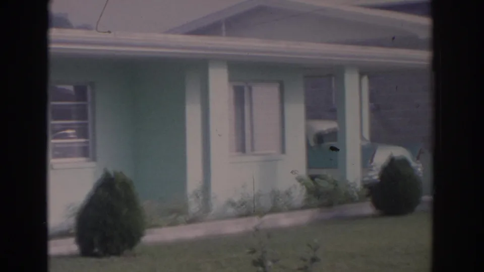1962 Florida 8mm frame showing the blue house exterior, white post, windows, lawn, shrub, and a partial view toward the carport.
