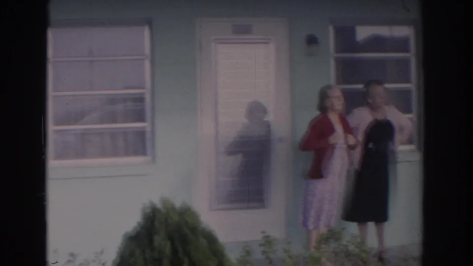 1962 Florida home-movie frame showing three women standing near the front door of a blue house with white trim and front landscaping.