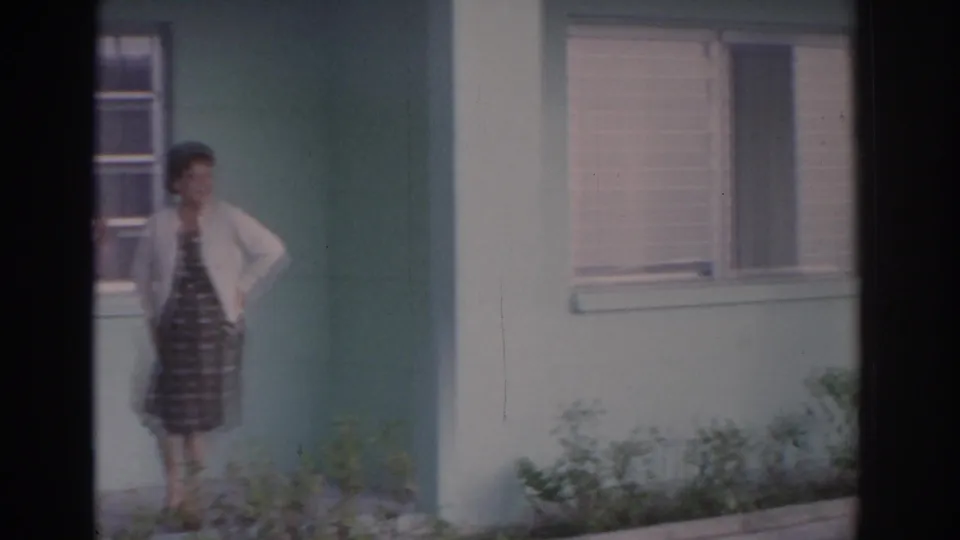 One adult woman in a patterned dress and white cardigan stands near the corner of a light green Florida house, with porch plants below.