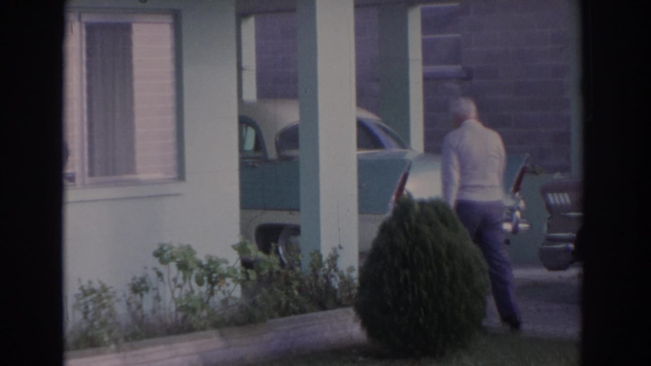 1962 Florida home-movie frame showing an elderly man walking outside a pale green house beside a parked late-1950s car.