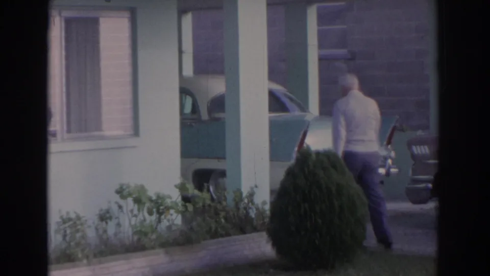 1962 Florida home-movie frame showing an elderly man walking outside a pale green house beside a parked late-1950s car.