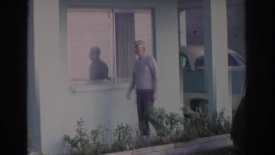 1962 Florida home-movie frame with an elderly man near a window, shrubs in the foreground, and a parked car beside the house.