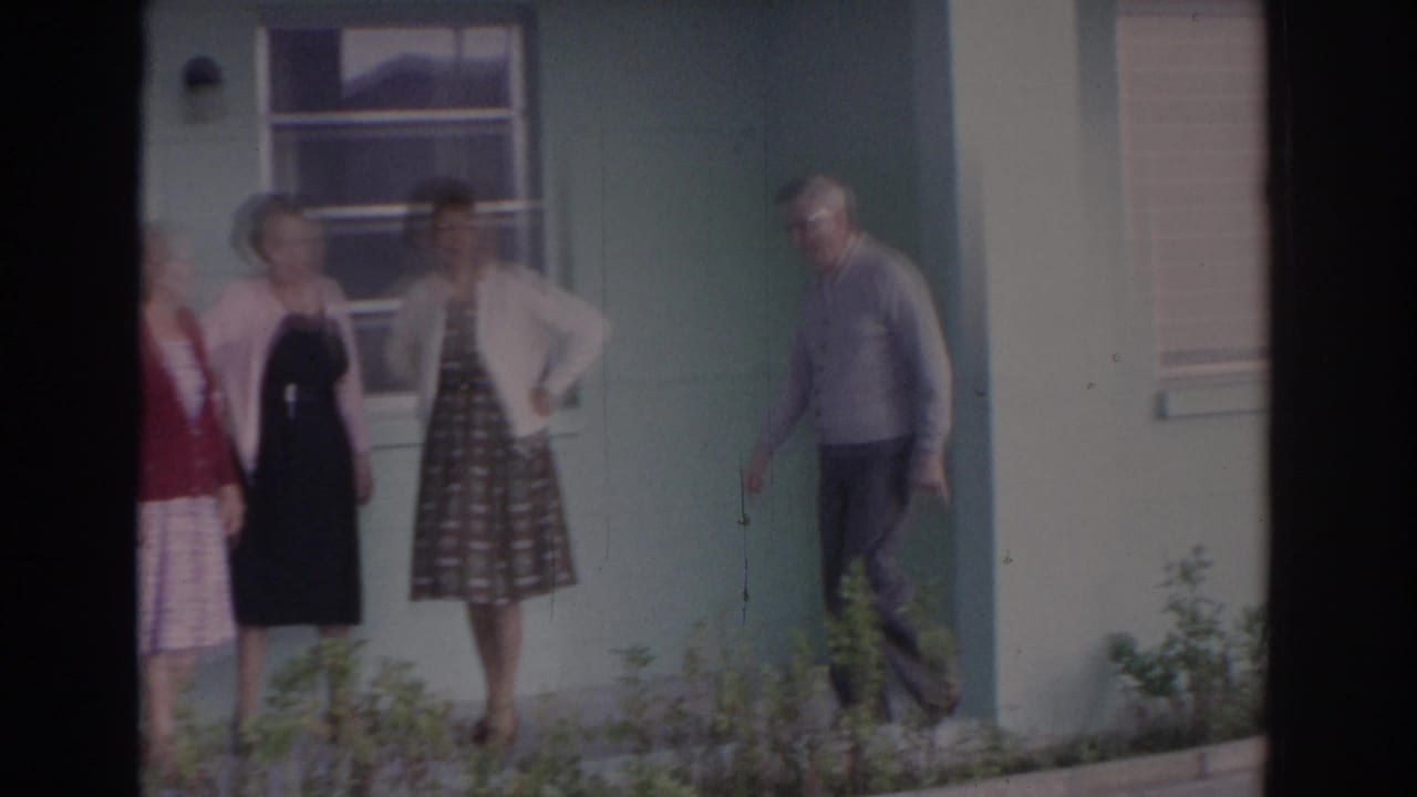 Older adults standing outside a pale green house near porch windows and shrubs.
