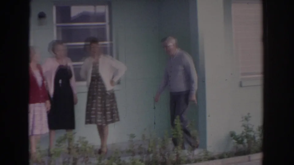 1962 Florida 8mm frame showing older adults in dresses and cardigans outside a pale green house with windows and shrubs.