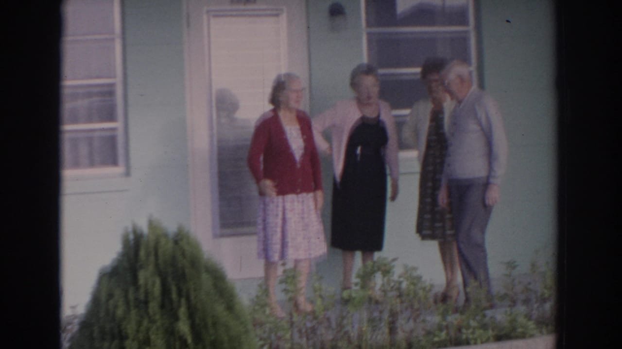 Four older adults standing together outside a pale house with windows and low shrubs.