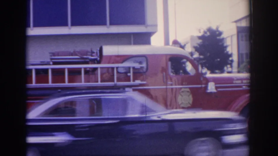 1962 Florida home-movie frame with a red fire truck parked by a building and a dark car passing across the foreground.