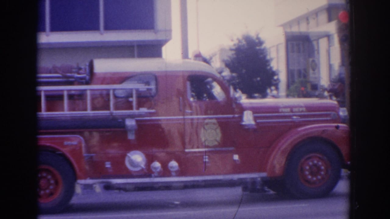 Red fire truck passing through a downtown intersection with buildings and traffic lights behind it.