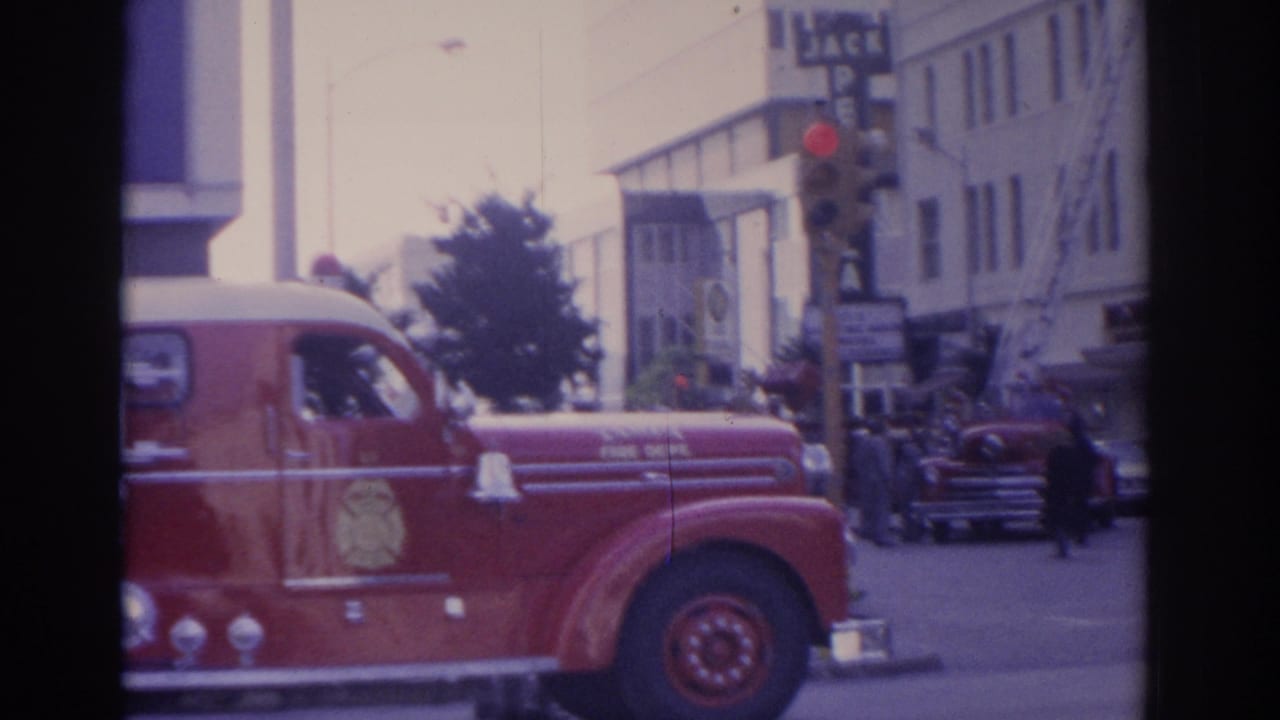 Red fire truck at an intersection near business signs, traffic signals, and older cars.