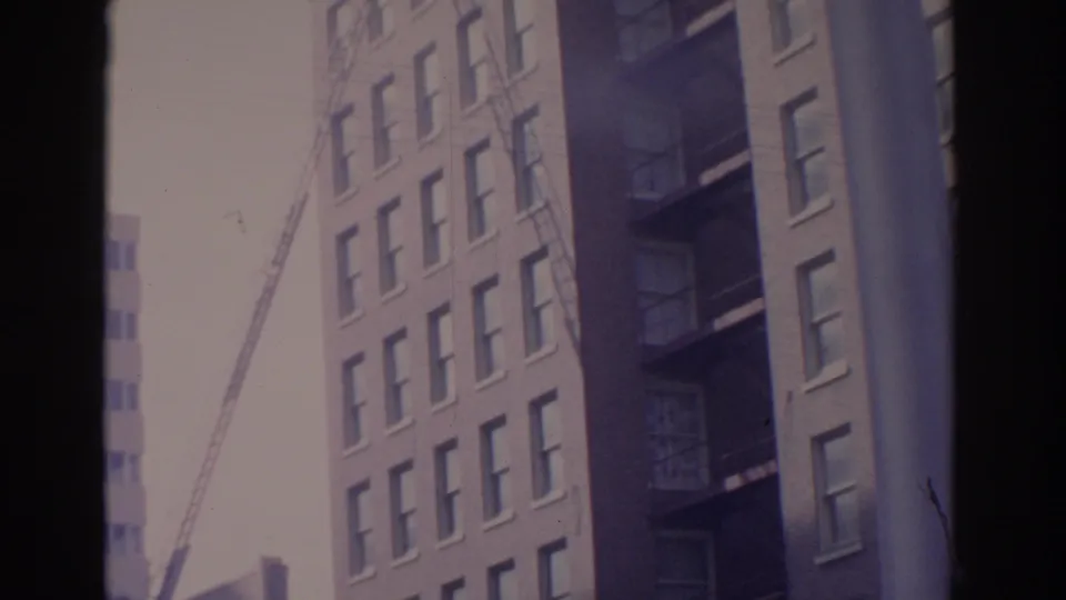 1962 Florida city frame showing a multi-story facade, fire-escape style balconies, and a ladder rising beside the building.