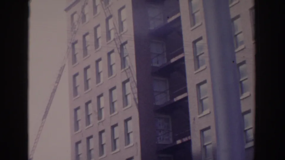 1962 Florida home-movie frame showing brick city buildings, exterior fire escapes, and a construction crane through dark frame edges.