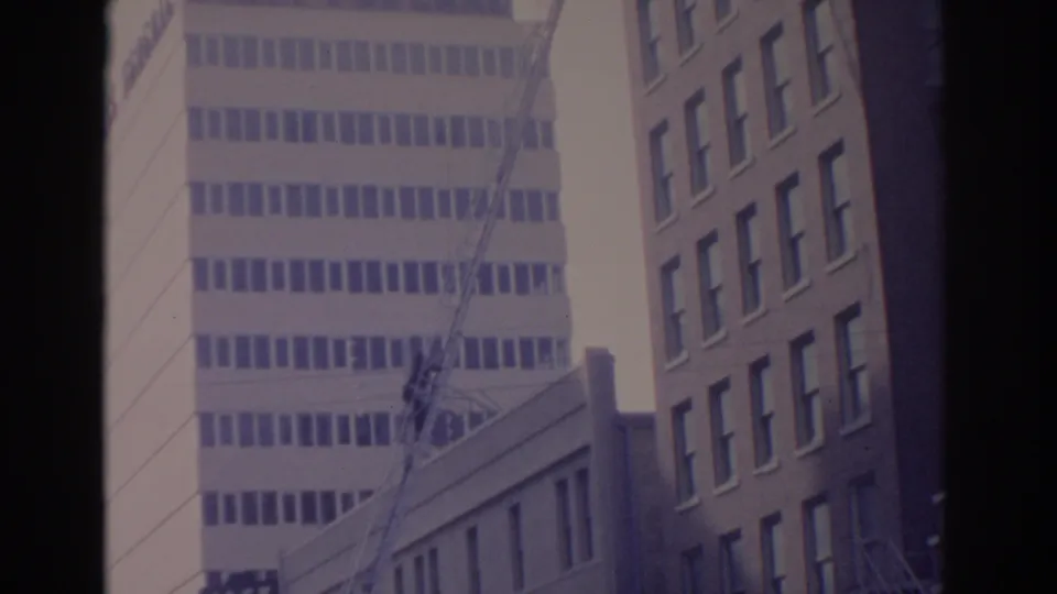 1962 Florida frame showing an urban construction crane near a pale office tower, with a brick building and dark film borders.