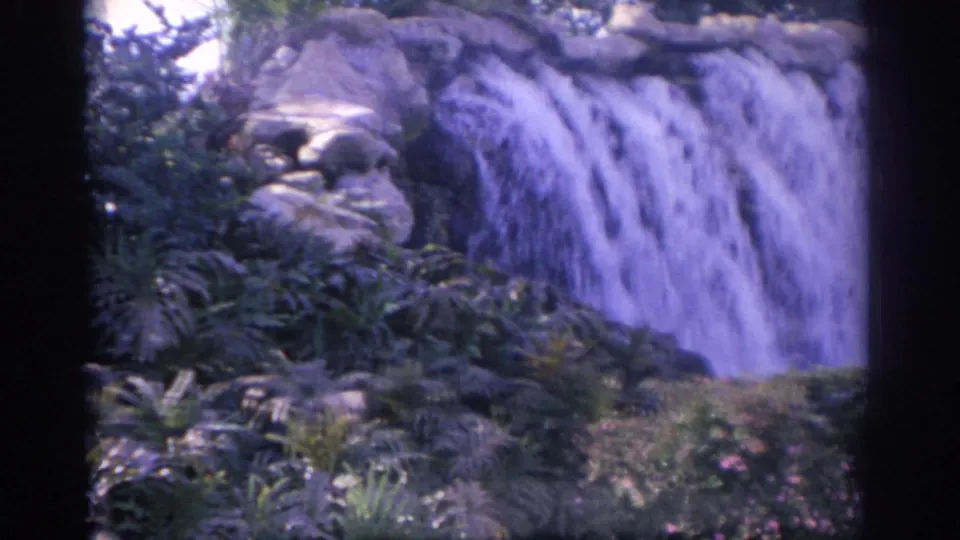 1962 Florida 8mm frame with water spilling over a rock ledge into a planted garden setting.