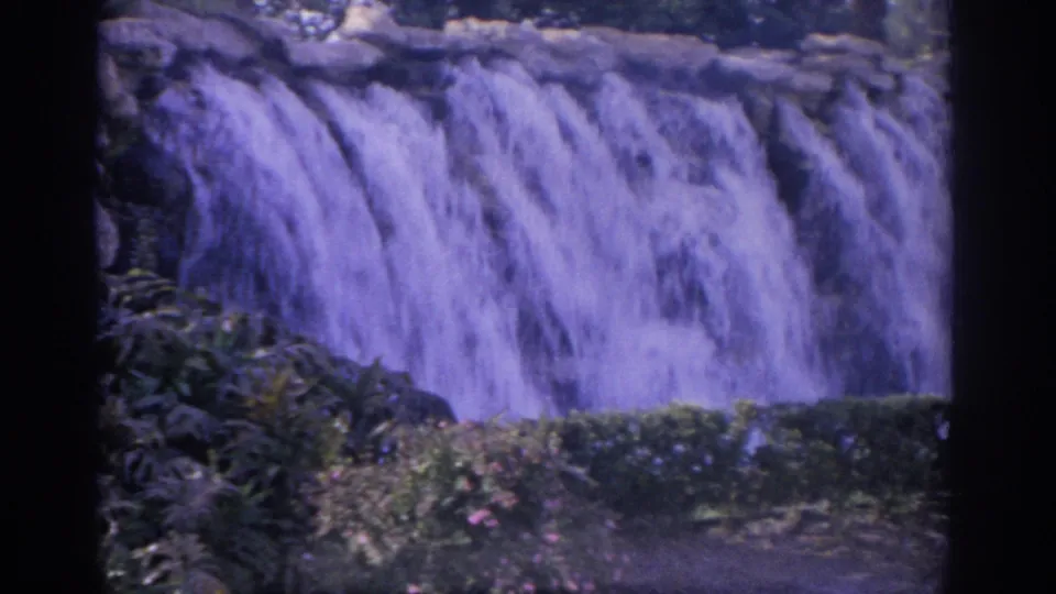 1962 Florida home-movie frame of a broad garden waterfall, with shrubs and rocks framing the lower edge.