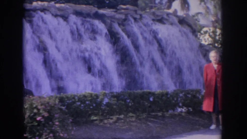1962 Florida 8mm frame showing an adult woman in a red coat standing beside a garden waterfall and low hedge.
