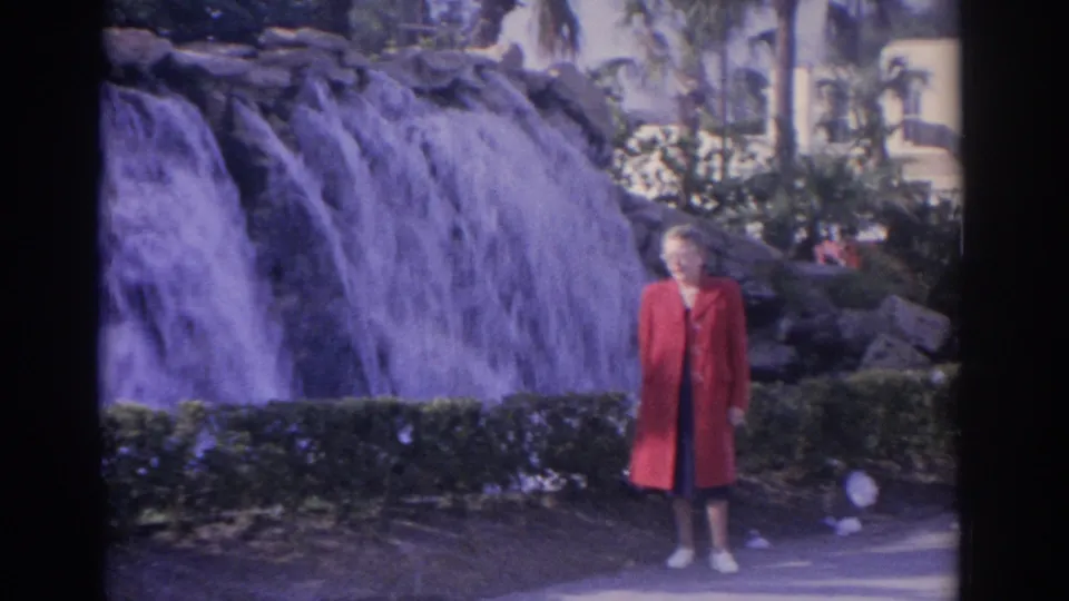 1962 Florida home-movie frame with an adult woman in a red coat standing on a paved path near the waterfall.
