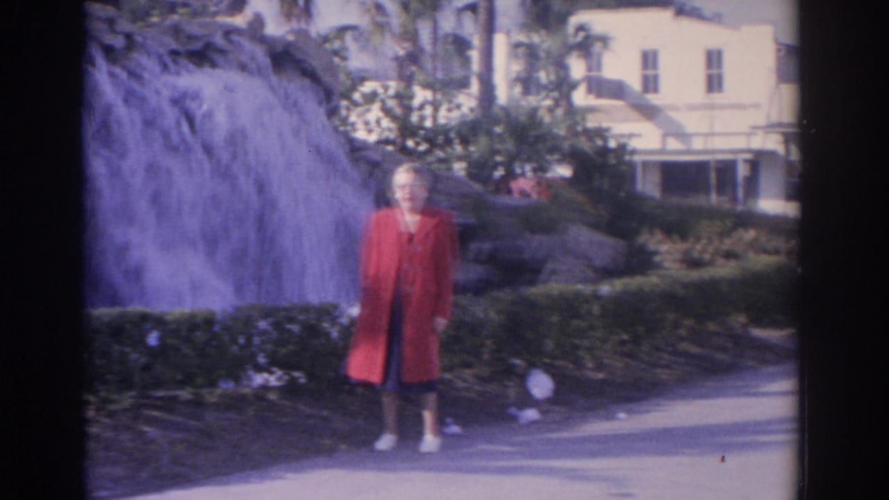 Woman in a red coat stands near a waterfall in a tropical garden.