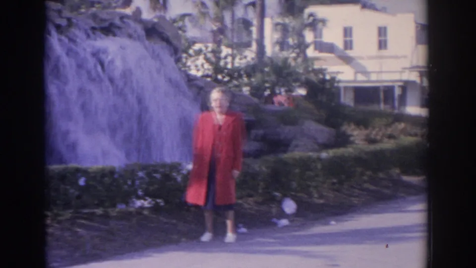 In 1962 Florida home-movie footage, a woman in a red coat stands by a cascading waterfall with palms and a pale building behind her.