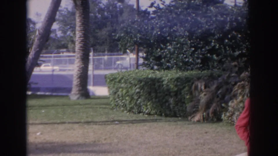 The camera pans across the Florida garden, showing lawn, hedges, palms, a road, and the red-coated woman partly cropped at frame edge.