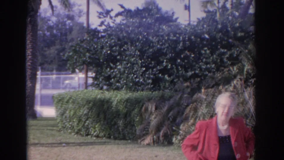 The woman in a red coat and glasses stands in a landscaped Florida garden, with trimmed hedges, palms, and roadside fencing behind her.