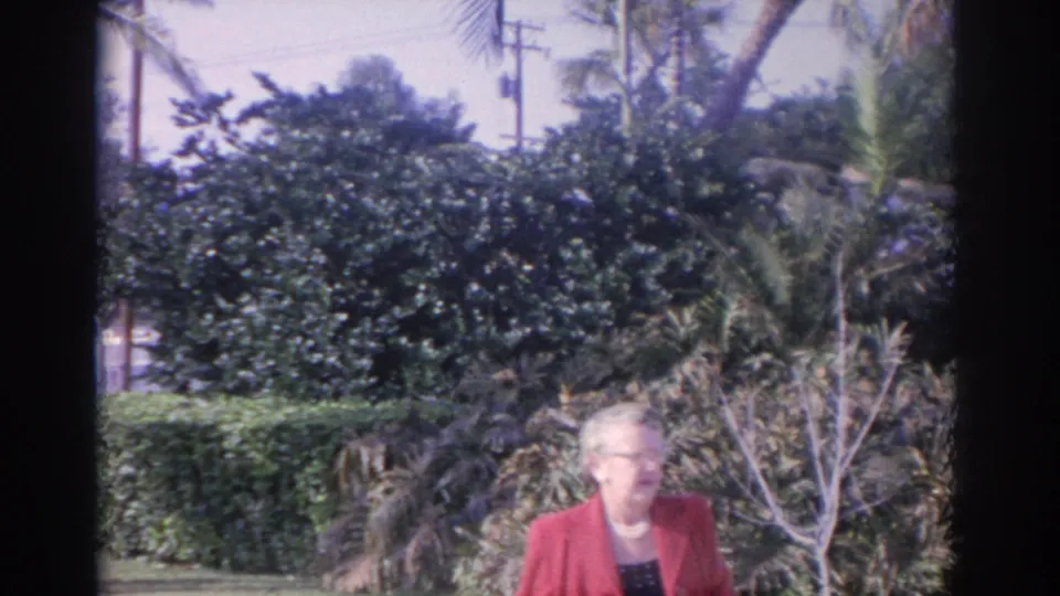 Close garden view of the woman in a red coat and glasses, with dense hedges, palms, and tropical plants in 1962 Florida footage.