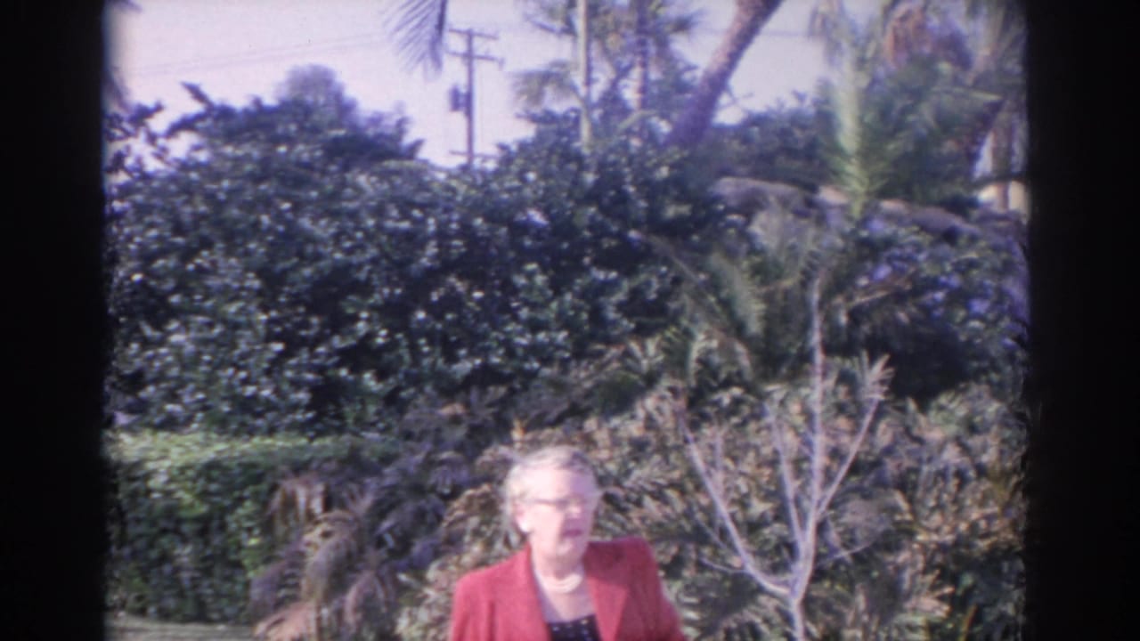 Older woman in a red jacket and glasses stands in a Florida tropical garden setting, consistent with the 1962 home-movie metadata.