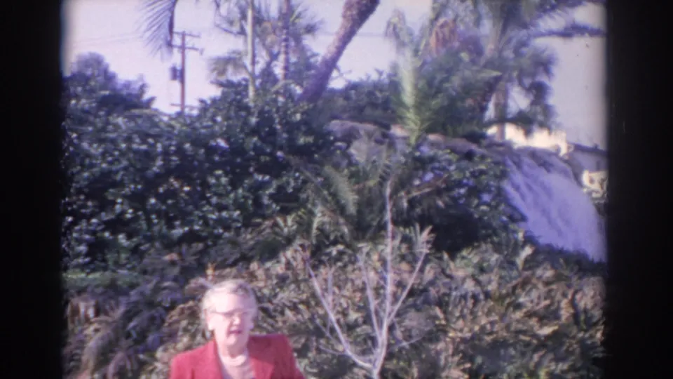 Older woman in red appears in a tropical Florida garden, with palms, thick shrubs, and a thatched-looking roofline in the background.