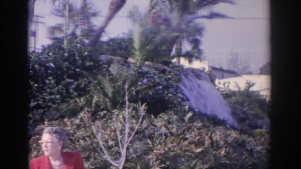 Older woman in a red jacket is partially framed at the lower edge as the camera shows tropical plants, palms, and nearby building details.
