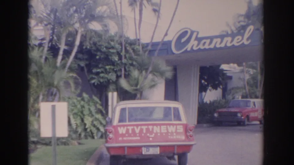 Red WTVT News station wagon is parked under a Channel sign at a Florida building entrance with palms and another red car visible.