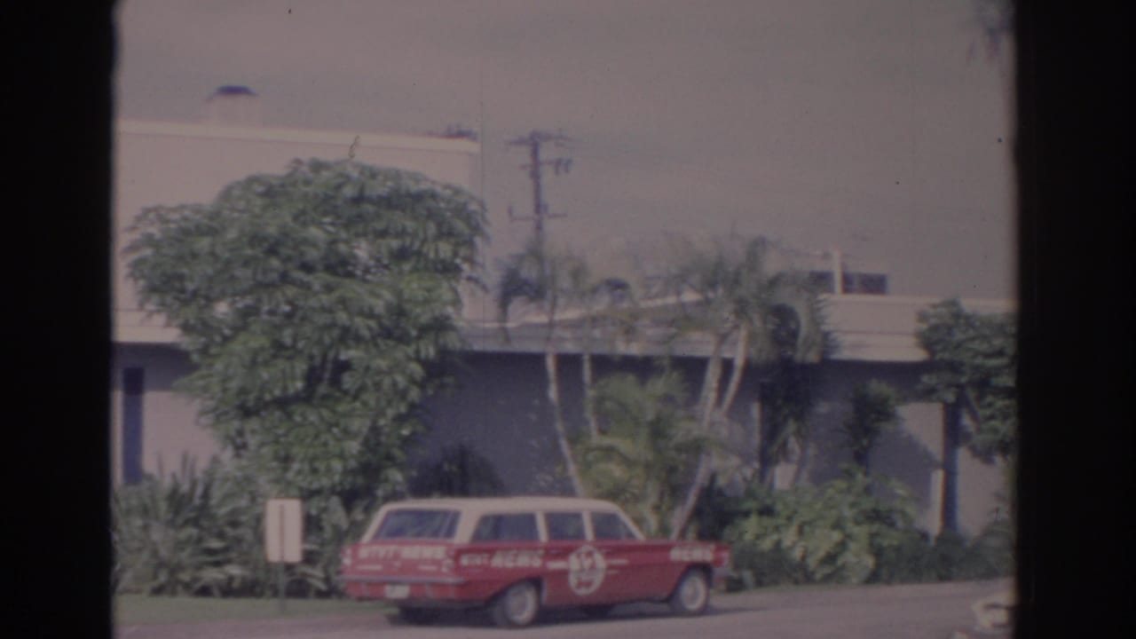 Red news wagon beside landscaping in front of a pale building.