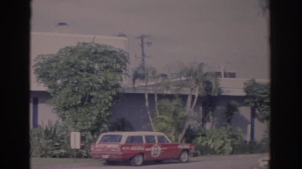 Red-and-white local news vehicle parked outside a Florida broadcast building, with palms and shrubs behind it.