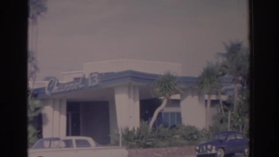 1962 Florida frame of the Channel 13 exterior with palm landscaping and parked mid-century cars near the entrance.