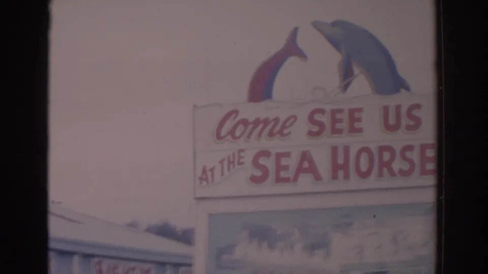 1962 Florida 8mm frame showing a Sea Horse invitation sign topped by dolphin-shaped figures, likely for a marine attraction.