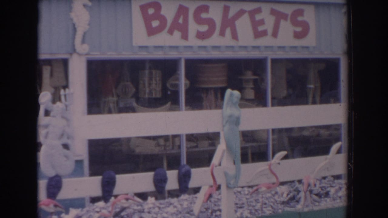 1962 Florida home-movie frame showing a light blue souvenir shop facade with BASKETS signage, craft displays, fence rails, and beach-themed decor.