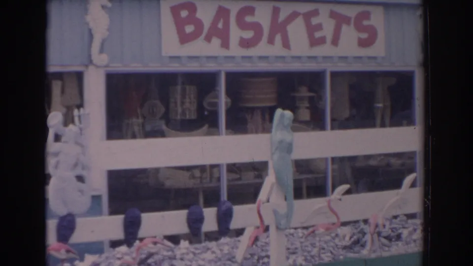 1962 Florida home-movie frame showing a light blue souvenir shop facade with BASKETS signage, craft displays, fence rails, and beach-themed decor.