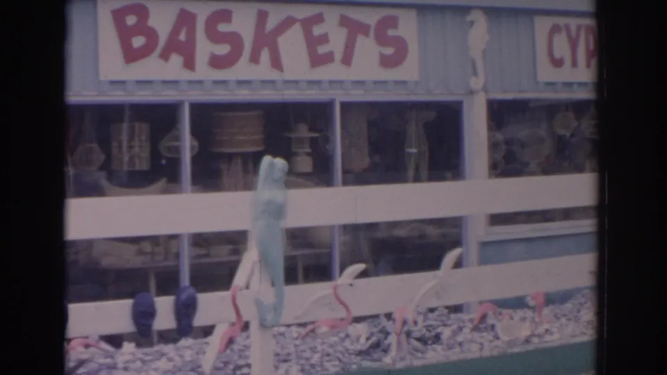1962 Florida storefront view with the BASKETS sign centered over display windows and a fenced row of flamingos, seahorses, and a mermaid figure.
