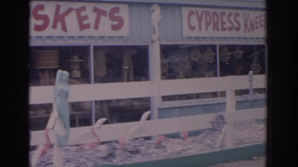 1962 Florida souvenir storefront as the camera reveals a CYPRESS KNEES sign beside partial BASKETS lettering, with window displays and beach decor below.