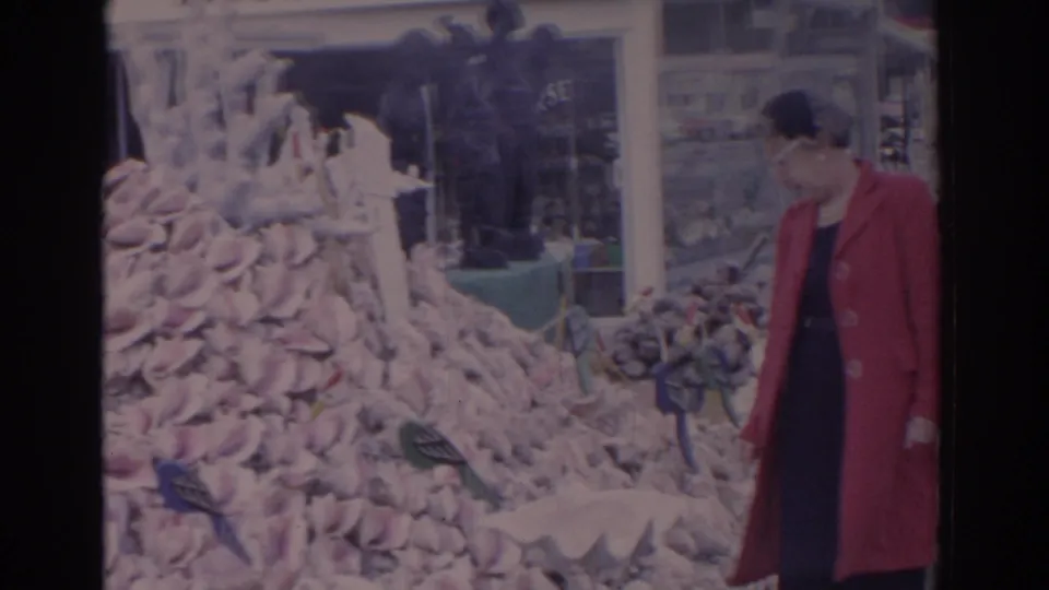A woman in a red coat and dark dress stands near piled conch shells at a Florida souvenir market in the stated 1962 footage.
