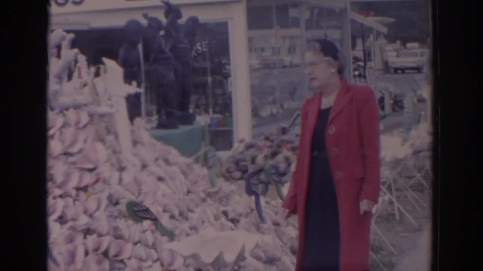 The woman in a red coat moves through the Florida market scene, with conch shells and souvenir displays filling the foreground.