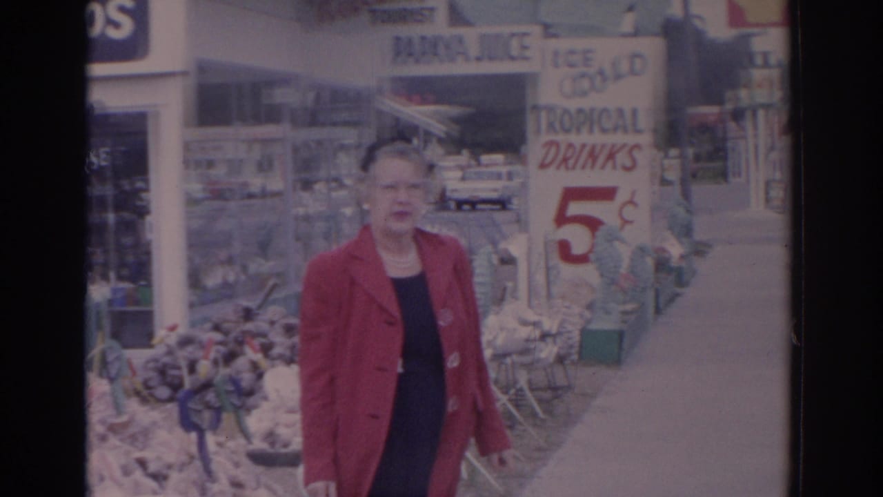 Woman in a red coat walks past a roadside drink stand with signs.