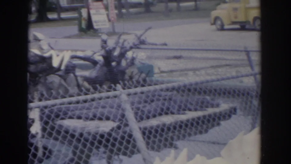 1962 Florida home-movie frame of alligator bags arranged behind chain-link fencing, with a yellow utility truck visible beyond the display.